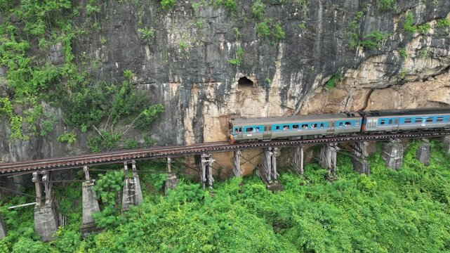 Train crossing bridge on famous Death Railway of Thailand. Aerial view from drone, dolly left. Popular tourist destination with scenic landscape and historic significance from World War Two.