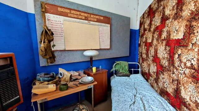 An abandoned Soviet-era room featuring a single bed, military gear, and a wall chart in Russian