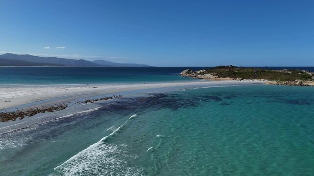 Waves of turquoise Indian Ocean reaching sandy beach in Tasmania. Sunny day with blue sky and mountains in background. Aerial wide shot.