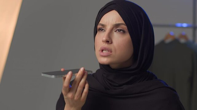 Angry displeased Islamic Muslim Arabian woman model lady speaking talk loudspeaker negotiate mobile phone smartphone call. Annoyed girl in hijab female record audio voice message voicemail in studio