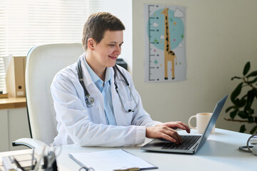 Portrait of doctor in white coat sitting at desk using laptop in bright office with giraffe poster on wall, stethoscope around neck, and notepad on table