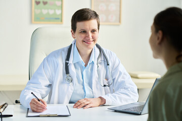 Portrait of senior female doctor wearing white coat and stethoscope, engaging in friendly conversation with patient while taking notes in office setting with medical charts in background