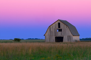 Obraz premium A weathered barn stands alone in a grassy field under a serene twilight sky transitioning from pink to blue hues