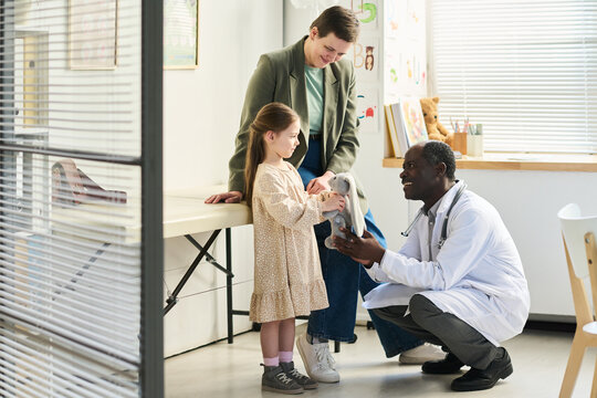 Smiling young girl visiting a pediatric clinic, interacting with a friendly doctor, holding medical equipment while her mother oversees situation