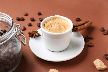Cup of hot espresso and jar with coffee beans on brown background