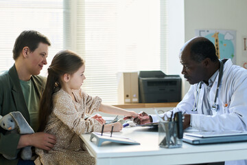 Child accompanied by parent during medical appointment, consulting with doctor in clinical office, holding a soft toy, engaging in conversation, displaying calm and attentive expressions