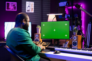 Young sound technician mixing music on console and piano with a greenscreen on pc display, home...