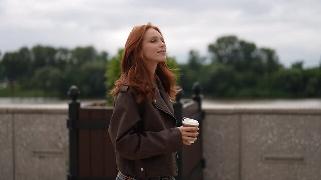 Side view tracking shot of charming woman with vibrant red hair strolling along city riverfront, enjoying peaceful moment with coffee, looking relaxed and carefree. Female in jacket enjoying free time