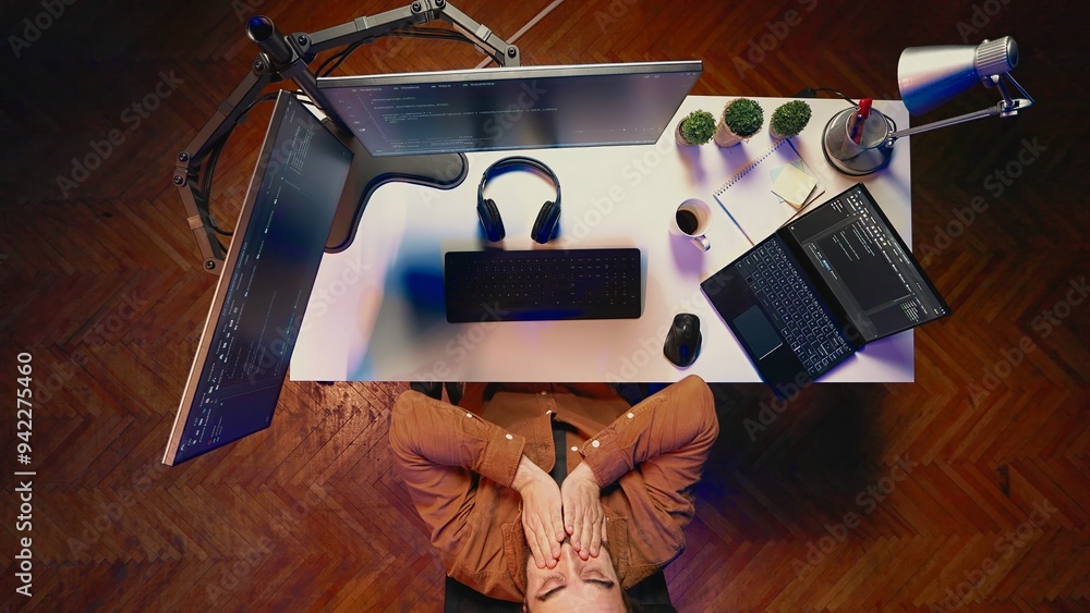 Poster top down view of fatigued man working overnight at home, rubbing eyes while taking break from coding