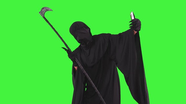 Grim reaper in Halloween costume holding a scythe taking a selfie with smartphone on a green screen