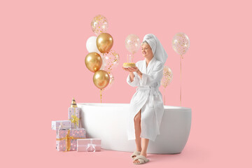 Happy young woman in bathrobe with birthday cake, gift boxes and balloons near bathtub on pink background