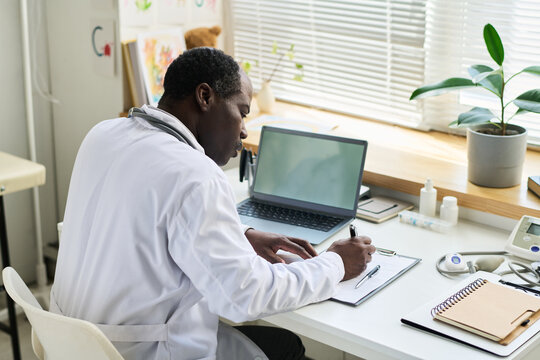Doctor in white lab coat writing notes at desk in modern office with laptop and medical tools surrounding. Large window providing natural light emphasizes workspace sophistication