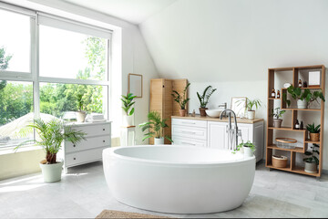 Bathtub, chest of drawers, folding screen and houseplants near window in light bathroom