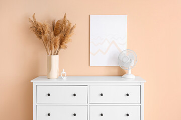 Modern electric fan on chest of drawers with pampas grass and picture on wall in room
