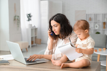 Cute little baby and young mother working with laptop while talking by mobile phone on maternity leave at home