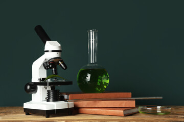 Modern microscope with chemical flask, petri dish and books on wooden table against green background