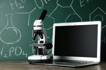 Modern microscope and laptop on wooden table near chalkboard with chemical formulas