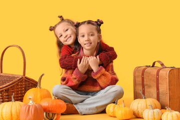 Cute little happy girls in autumn clothes with pumpkins, wicker basket and suitcase sitting on yellow background