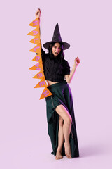 Beautiful young happy woman dressed as witch for Halloween with garland on lilac background