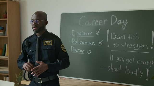 Medium slowmo shot of cheerful African American policeman in uniform speaking to students about personal safety measures, pointing to rules on blackboard, while attending career day at school
