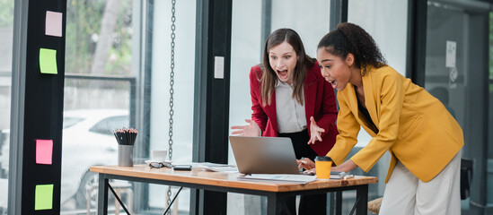 Two enthusiastic businesswomen working together on a project in a modern office, expressing excitement while looking at a laptop.