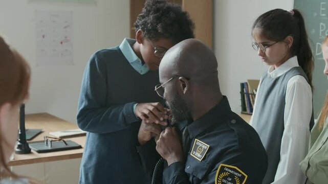 Medium shot of young black security officer or bodyguard in uniform sitting in classroom, chatting to multiethnic boy and girl students at school career day, letting play with portable shoulder radio