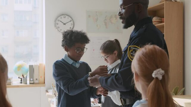 Medium shot of smiling black policeman in uniform chatting with class of diverse school students on career day, letting kids play with torch and handcuffs, talking about profession