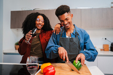 African American couple is cooking vegan food in their apartment kitchen while enjoying a glass of red wine. Two young people are cutting vegetables to prepare a healthy vegetarian meal at home.