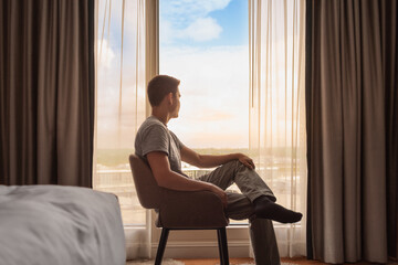 Thoughtful young man looking out his bedroom hotel window at the beautiful view