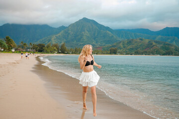 Cheerful blonde in a black top and white skirt poses on the beach against the background of green mountains, Kauai, Hawaii