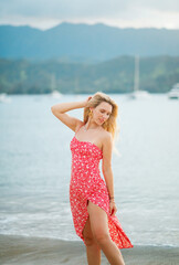 A beautiful woman in a red dress with a print is posing on the beach at sunset, Hawaii