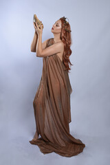 Full length portrait of beautiful female model wearing silken toga robes with golden crown, historical fantasy greek goddess aphrodite costume, holding seashell skull. Isolated on studio background