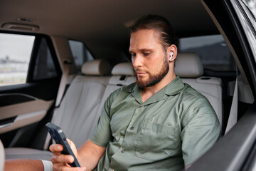 Young man with earbuds using a smartphone in the backseat of a car