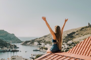 Woman sits on rooftop with outstretched arms, enjoys town view and sea mountains. Peaceful rooftop relaxation. Below her, there is a town with several boats visible in the water