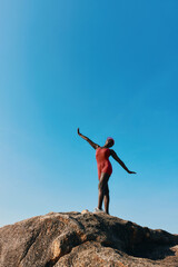 A swimmer in a red swimsuit poses on a rock against a clear blue sky, showcasing strength and...