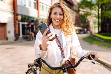 A young woman checks her phone while leaning on  bicycle in the city during a sunny afternoon stroll. Selfie time. People, leisure, blogging and lifestyle