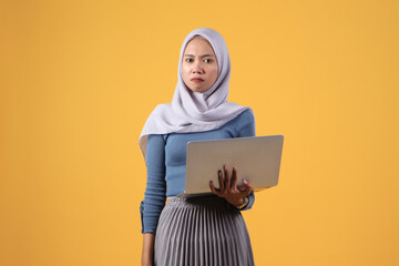 asian indonesian muslim woman holding laptop computer with angry face expression on isolated yellow background