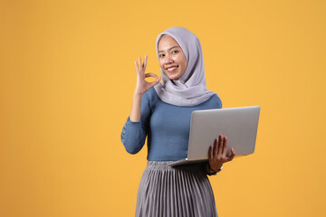 happy asian indonesian muslim woman holding laptop computer giving ok finger sign on isolated yellow background