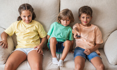 Three Happy And Smiling Siblings Or Friends, Preschool And Elementary Age, With Positive Emotions, Enjoy Playing Or Watching Television While Sitting On The Sofa At Home.