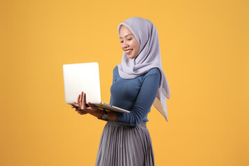 happy asian indonesian muslim woman holding laptop computer on isolated yellow background
