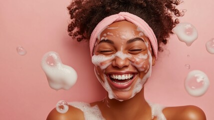 A happy woman with cream/soap, isolated on a pastel pink background. A beauty portrait of a smiling African American girl, smiling while washing her face during her morning skincare routine.