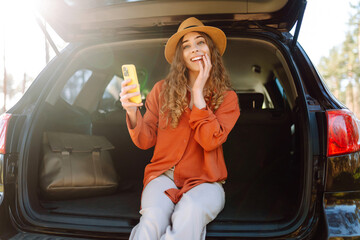 A young woman enjoys a sunny day by the car while using her smartphone, wearing stylish sunglasses...