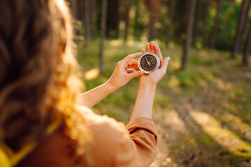 A hiker with curly hair holds a compass in their hands, carefully navigating through a sunlit forest filled with tall trees, greenery, and warm afternoon light. © maxbelchenko