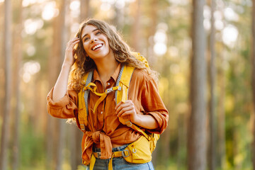 Happy young woman with curly hair walks along a forest path, wearing a yellow hiking backpack surrounded by tall trees and warm sunlight. Trips. Active lifestyle. Hiking.