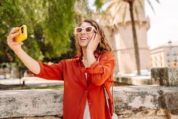 Cheerful woman taking a selfie in stylish sunglasses while strolling down a sunlit city street...