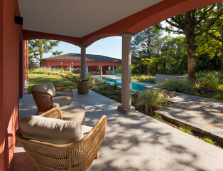 Outdoor patio of a villa, furnished with two outdoor armchairs and a small table, perfect for relaxing. In the background you can see the outbuilding and the swimming pool. Sunny day, no one inside