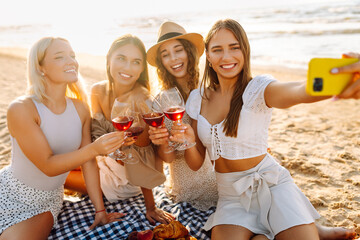Four beautiful women friends enjoying a beach gathering while taking selfies and sipping drinks...