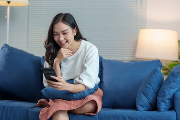 Fototapeta premium Happy and relaxed Asian woman sitting on sofa on weekend using smartphone to communicate, talk, text, chat online, watch wireless internet on device. Shopping online from your home office.