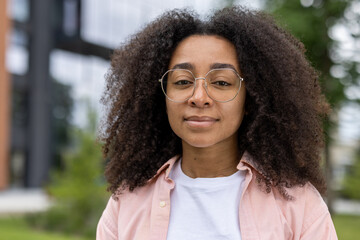 Portrait of confident young woman with curly hair and round glasses outdoors. She wears a pink shirt and white t-shirt, exuding calm and self-assurance. Urban background adds modern feel.