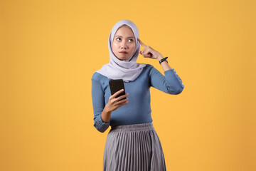 pensive asian indonesian muslim women holding smart phone with thinking gesture on isolated yellow background